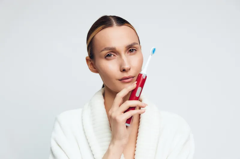 Young woman in white bathrobe holds red electric toothbrush near face looks at camera with calm expression isolated on white background personal hygiene concept.