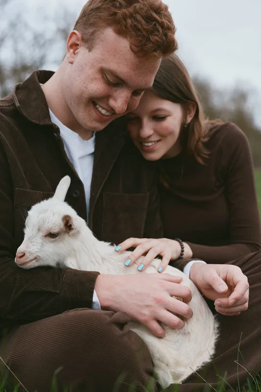 Lifestyle couple outdoors with a playful white goat kid, sitting in a grassy field, sharing a warm, affectionate moment, soft natural light, rustic charm, simple happiness