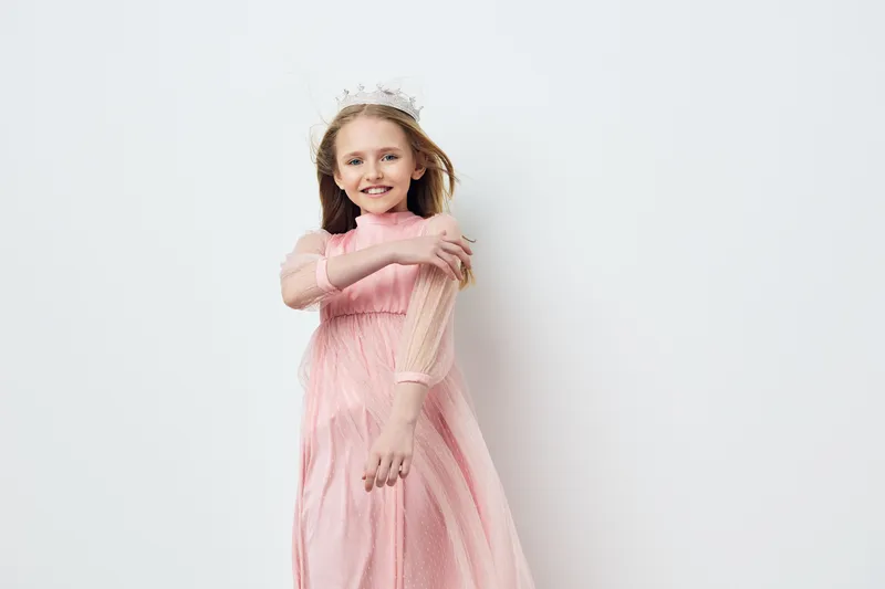 Cute girl in a pink dress with a crown, smiling joyfully against a light background, embodying childhood happiness and innocence in a studio setting