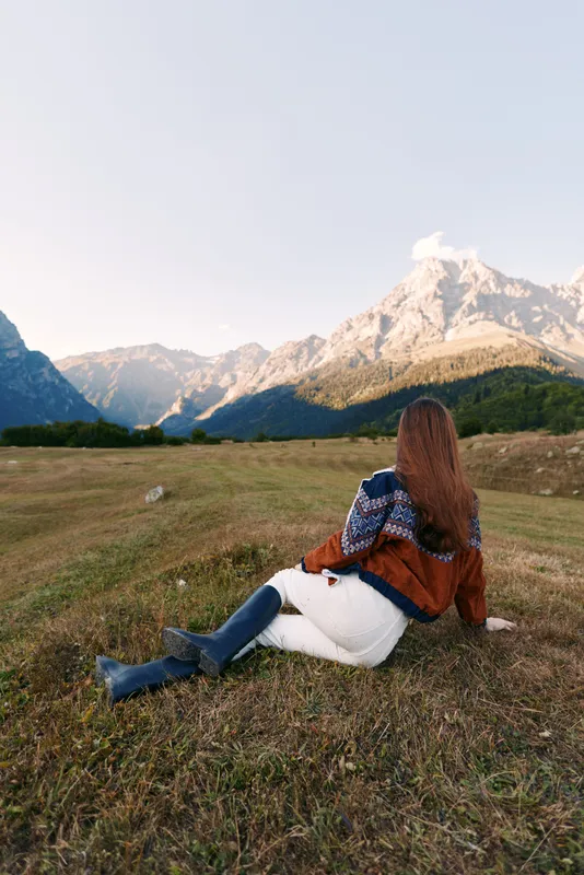 Woman sitting in a meadow gazing at majestic mountains, nature landscape during autumn travel and solitude, cozy sweater and boots on grassy valley with serene outdoor scenery and calm.