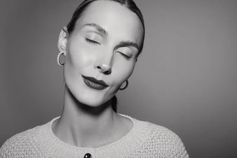 Close-up of a young Caucasian woman with fair skin and closed eyes, wearing hoop earrings and a knit sweater. Soft lighting highlights her serene, calm expression in monochrome.