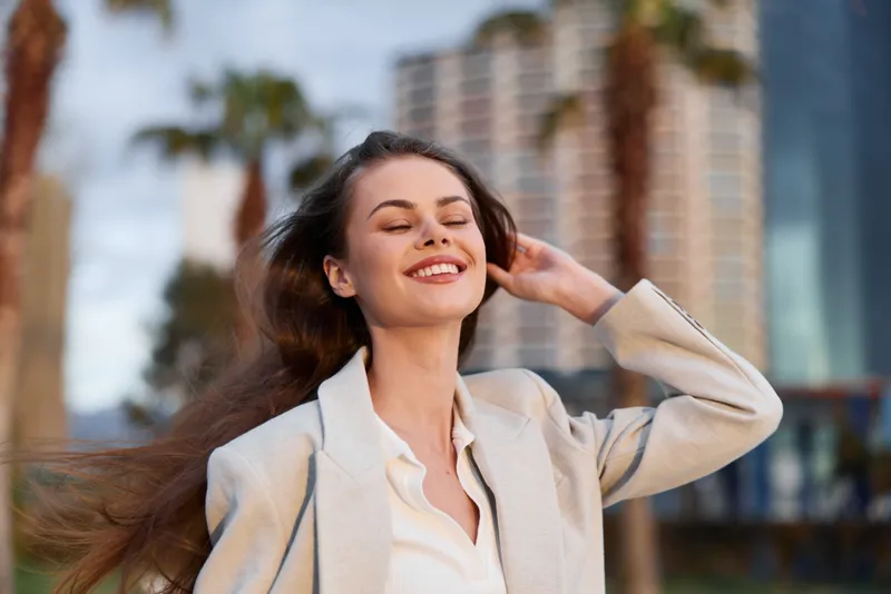 happy woman smiling outside in beige coat with long brown hair and urban background in warm daylight fashion lifestyle
