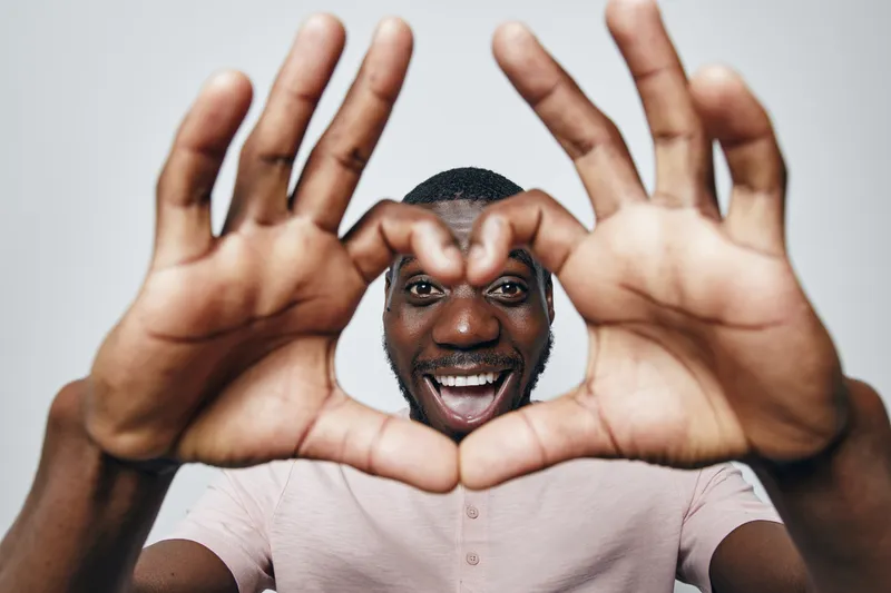 African American man with dark skin smiling and making heart shape with hands close to camera. Portrait of happy male showing love gesture with fingers on white background.