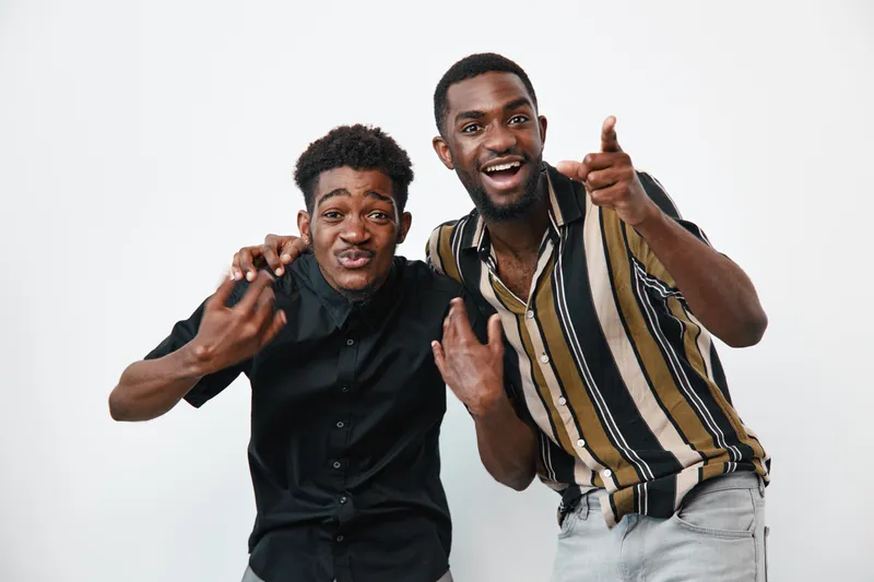 Two young black men with dark skin posing together in casual clothing on white background. Friends smiling and making hand gestures for fun and connection in studio.