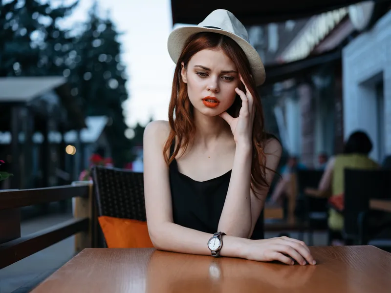 Женщина в белой шляпе и черном платье сидит за столиком в уличном кафе