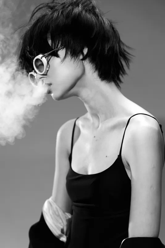 Black and white portrait of a young woman wearing sunglasses and exhaling smoke in a minimalist style.
