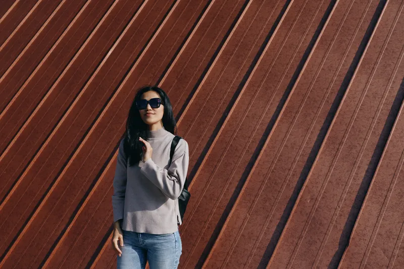 Woman in sunglasses wearing a grey sweater and jeans stands beside a diagonal wooden wall, casual fashion, outdoor urban scene with a relaxed pose