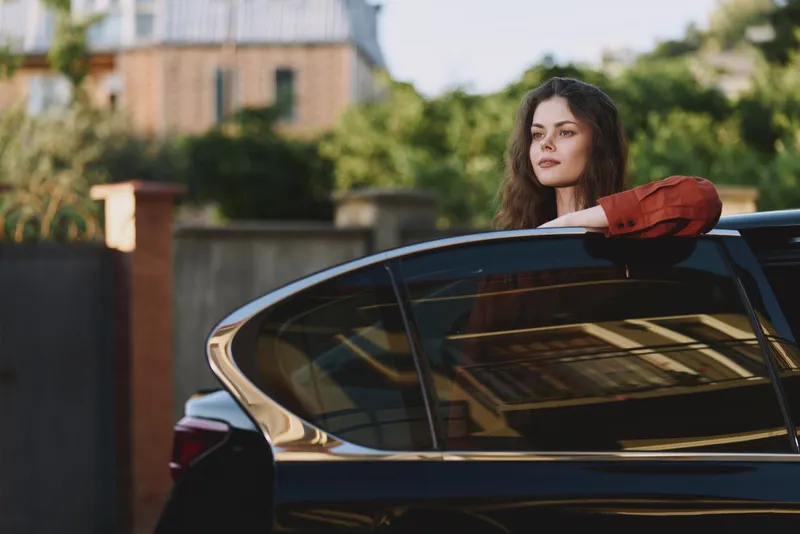 Confident woman with long hair leaning on a sleek car in an outdoor urban scene, highlighting a real life lifestyle, casual fashion, and relaxed self expression