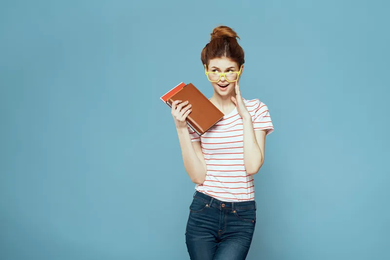 Студентка с книгами в руках на голубом фоне, в жёлтых очках, с модельной причёской, кадрированный вид