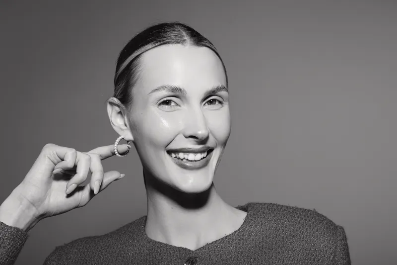 Smiling Caucasian woman with fair skin and slicked hair touches her ear wearing hoop earrings. Studio portrait with soft lighting and grey background. Concept of happiness and elegance.