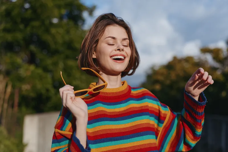 Young woman in her mid 20s smiling woman outdoors, joyful and carefree, wearing a colorful striped sweater and holding yellow sunglasses, casual outfit lifestyle portrait in a sunny park.