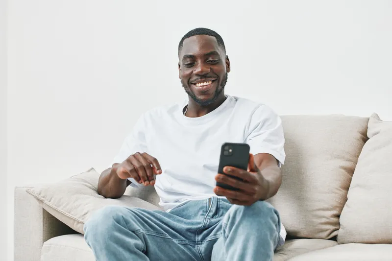 Happy young Black man smiling while using smartphone on a couch in a bright living room, showcasing modern lifestyle and technology engagement
