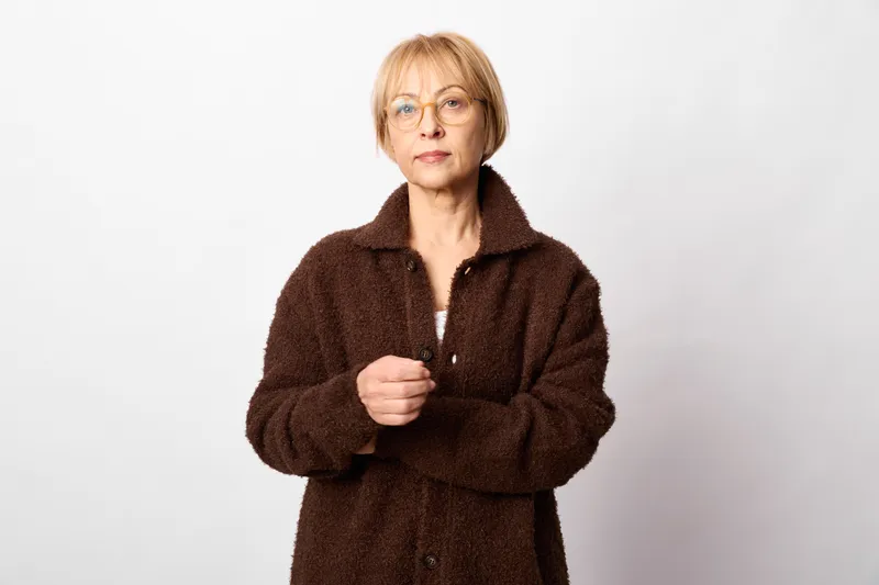 woman senior portrait coat glasses studio white winter fashion calm, mature female looks at camera, indoor overcoat, hands holding collar, neutral light