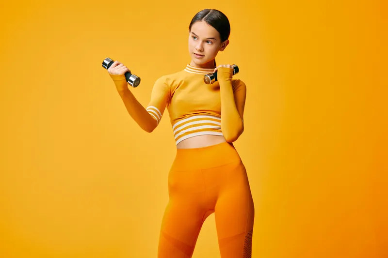 Athletic woman in bright orange fitness outfit lifting light dumbbells, posing with focused expression and strong posture in a minimalist studio with warm yellow background.