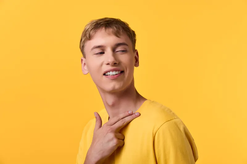 Smiling man in a yellow shirt poses with a casual neck gesture against a bright yellow backdrop, conveying warmth, style, and a cheerful everyday lifestyle moment.
