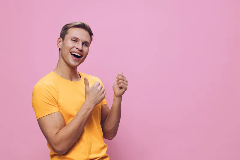 Cheerful smiling man in casual yellow t-shirt expressing happiness and excitement, gesturing with fists raised, against pink background. People lifestyle concept, positive emotion, youthful male, joyf
