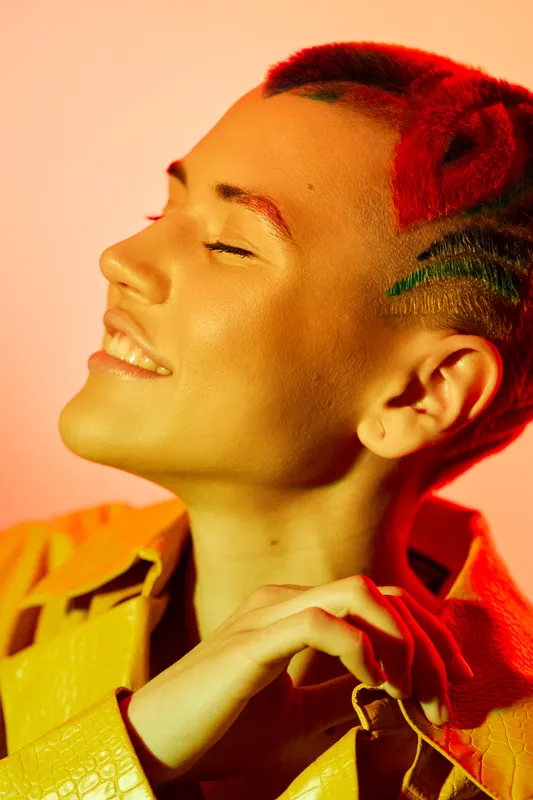Smiling woman with colorful hair and vibrant makeup, enjoying warm lighting and wearing a bright yellow jacket, expressing joy and confident style.