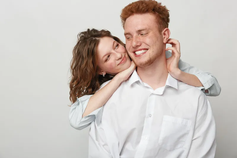 Happy young couple in white shirts embraces and smiles, showing affection and love. Studio portrait on light background. Concept of relationships, romance, and positive emotions.
