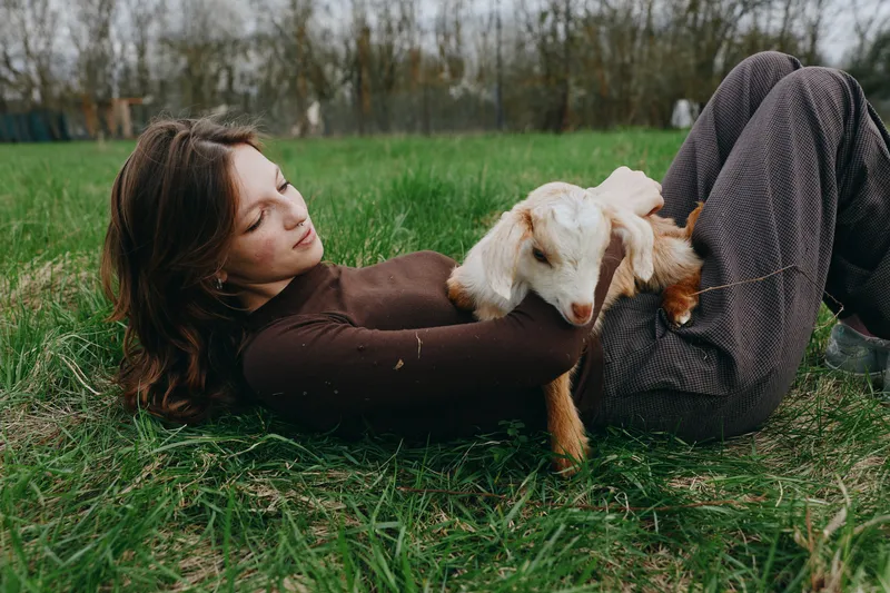 Young woman lies on the grass holding a baby goat in her arms enjoying a peaceful moment in a natural outdoor setting during autumn season.
