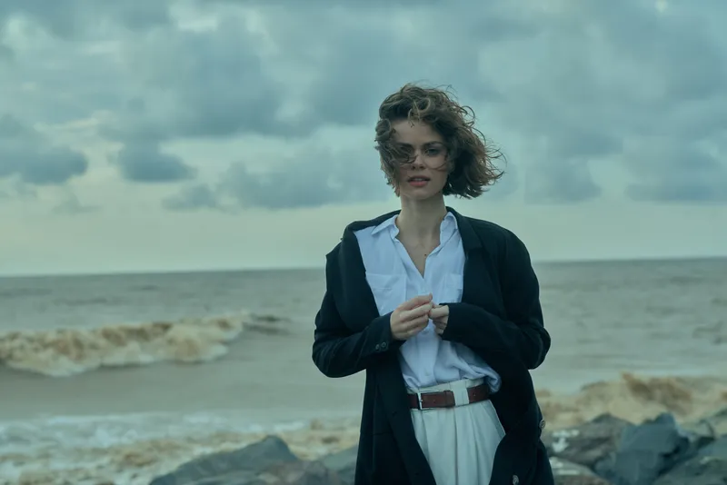 Woman in blazer and white shirt stands by a windy sea, dramatic skies create a moody coastal fashion scene against the ocean backdrop