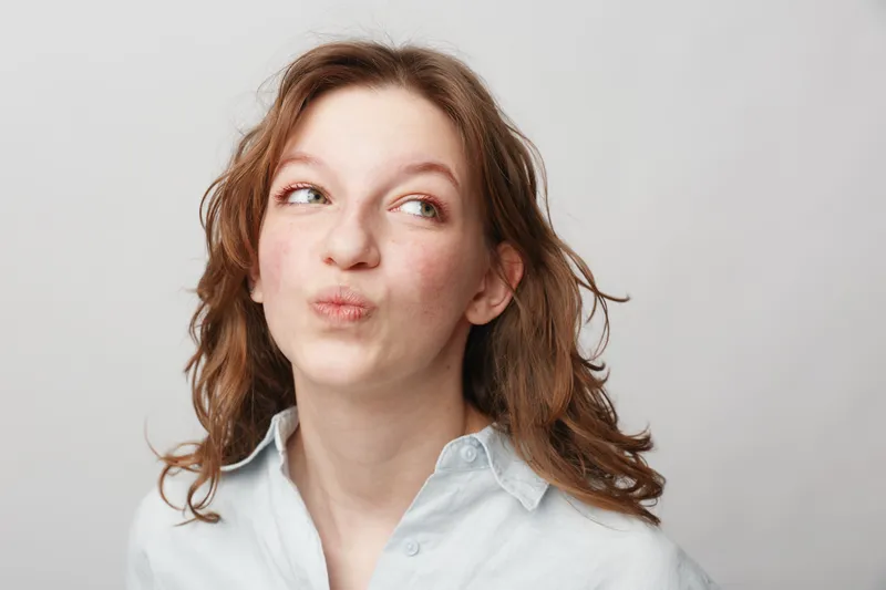 Young woman with curly hair looks sideways with a thoughtful expression wearing a light shirt on a plain background. Concept of contemplation, curiosity, and casual portrait.