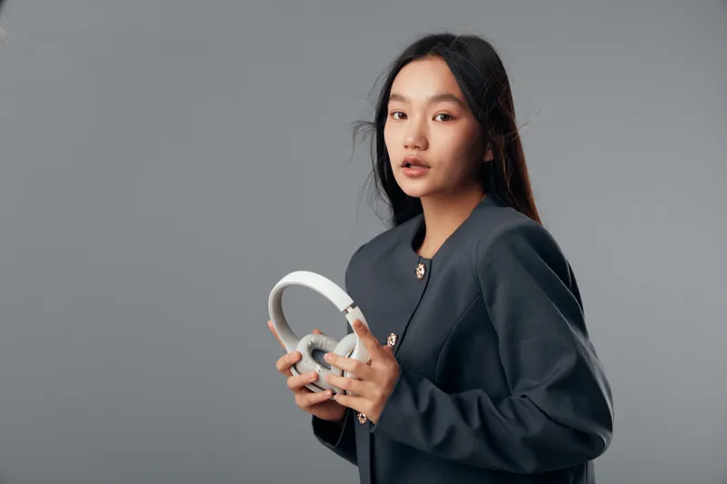 Young woman holding white headphones in hands looks at camera with slight expression on face dressed in dark jacket against grey background in studio portrait.
