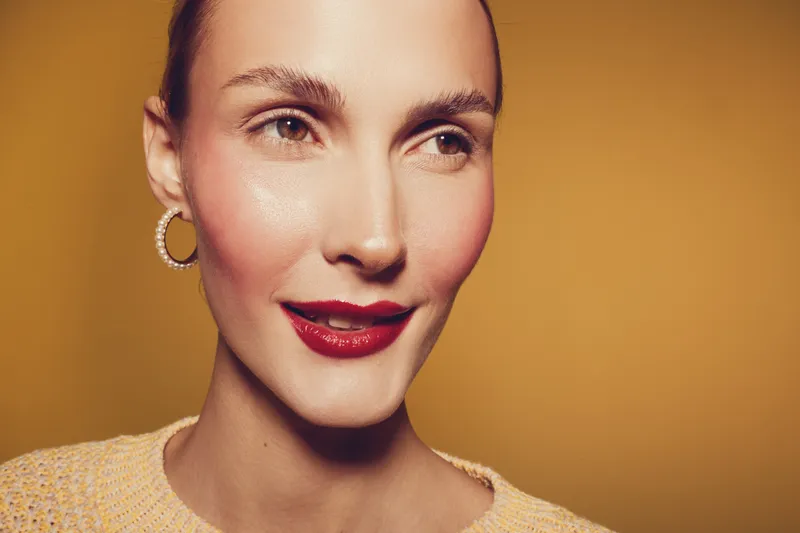 Close-up portrait of a smiling woman with red lipstick and hoop earrings on yellow background. Concept of beauty, fashion, makeup, and confident female model with natural skin.