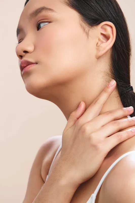 Asian woman appears in a simple studio portrait, calm and graceful, with a delicate touch along the neck, conveying poise, confidence, and refined style in a soft, balanced setting.