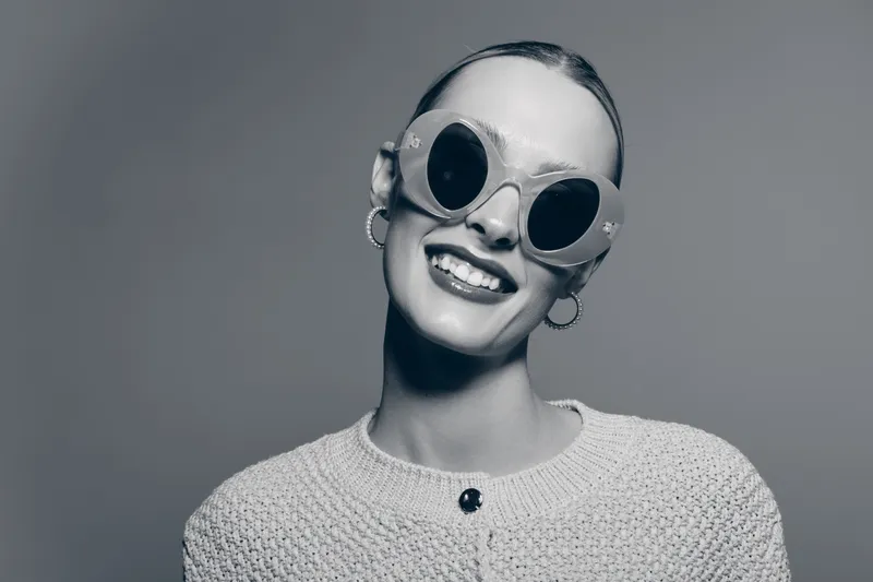 Smiling white woman with light skin wearing large round sunglasses and hoop earrings in studio. Concept of fashion, joy, and retro style.