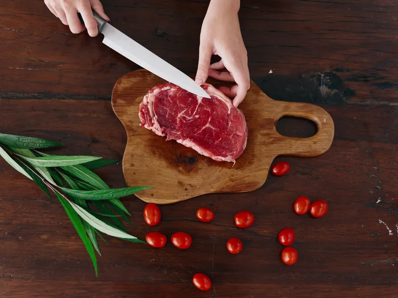 Женщина с ножом в руке нарезает свежее стейковое мясо для жарки на кухне; на столе соль, перец и другие специи, красные помидоры черри и зелень, идет подготовка к ужину