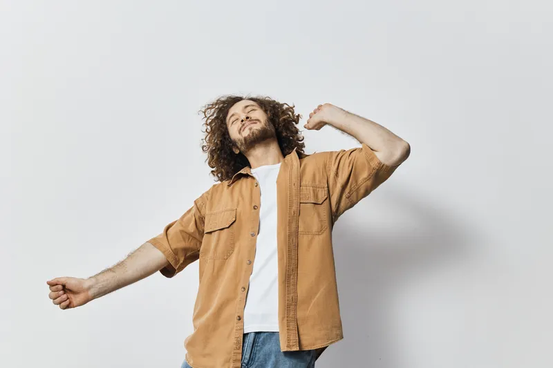 Young man with curly hair in a relaxed pose wearing a casual outfit, expressing happiness against a light neutral background, perfect for lifestyle and fashion concepts
