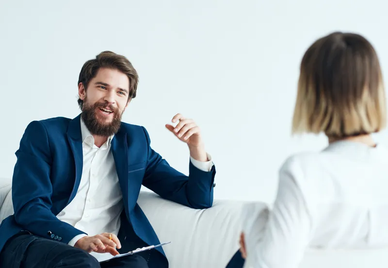 Мужчина в классическом костюме сидит на диване в помещении с документами, а рядом женщина