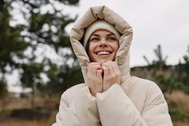 Woman in a warm hooded coat outdoors, smiling in a misty natural setting, capturing cozy winter mood and optimistic outdoor vibe for lifestyle photography.