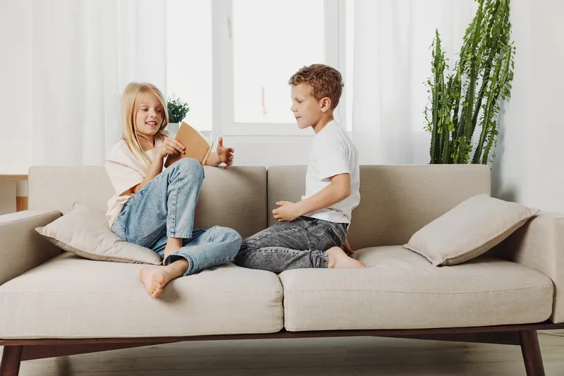 Two children sitting barefoot on beige sofa in bright living room, smiling and playing together. Concept of childhood, friendship, family bonding, and indoor leisure activity.