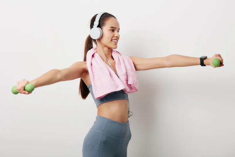 Young woman in athletic activewear doing lateral dumbbell raises, wearing headphones and smartwatch, pink towel draped around neck, smiling against a clean white studio background.