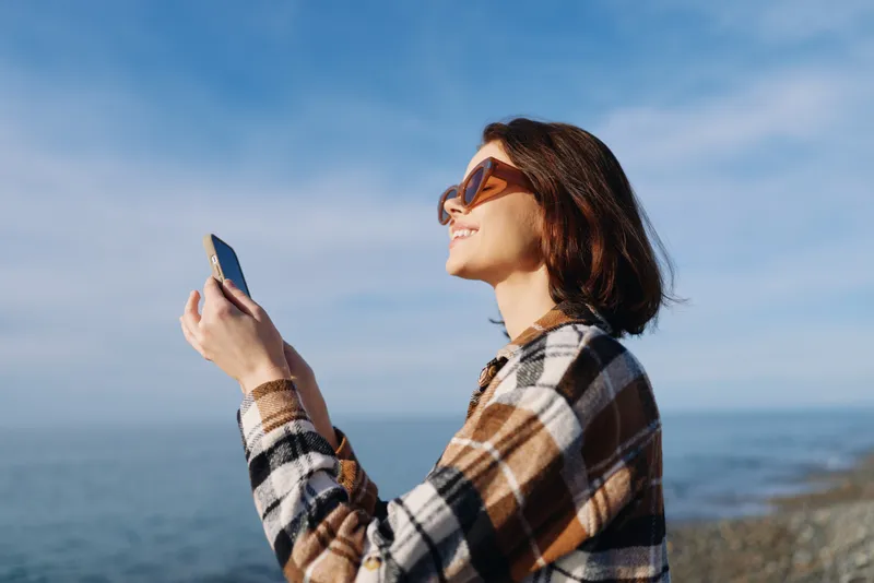 woman, smartphone, outdoor, sea, beach, sunglasses, plaid shirt, smiling portrait enjoying sunny weather while capturing moments by the water with a mobile device and relaxed vibe