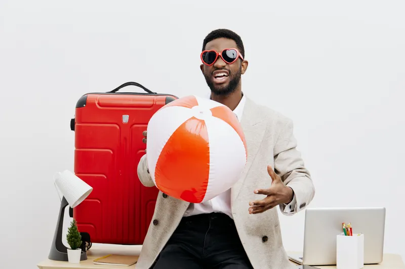Vibrant young man with dark skin wearing stylish sunglasses and light blazer, holding a colorful beach ball, standing in front of a travel suitcase and desk, cheerful vibe. People lifestyle concept, t