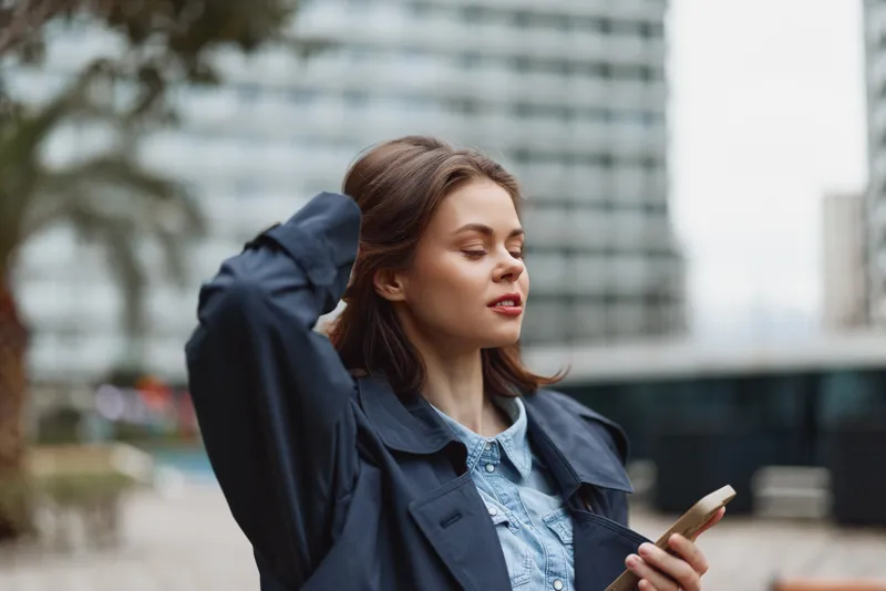 Young woman with closed eyes holding smartphone in city street, wearing blue coat and denim shirt. Concept of relaxation, technology, urban lifestyle, and casual fashion.