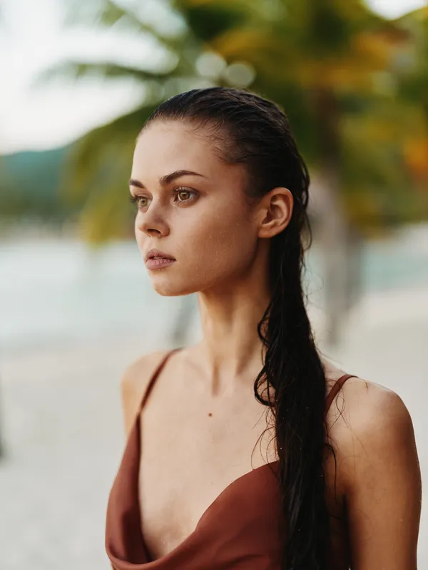 Portrait of a pensive woman on a sunlit beach, with wet hair and a warm swimsuit; a calm sea forms the backdrop as a thoughtful expression centers the frame.