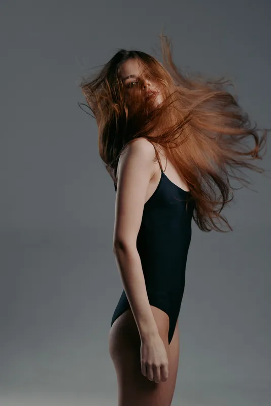 young woman in black bodysuit with flowing red hair against gray background