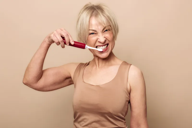 Middle aged woman with blonde hair brushes teeth with electric toothbrush in beige tank top on neutral background. Concept for dental care, hygiene, and healthy lifestyle.