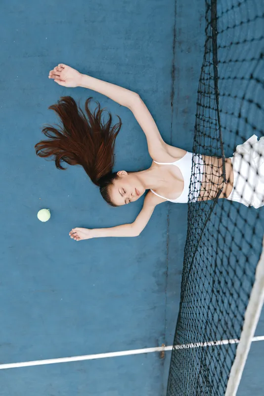 Young woman lying on a tennis court with long hair and a tennis ball, showcasing relaxation and sportiness in a vibrant blue setting