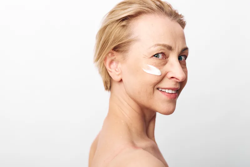 Mature woman with light skin applies white face cream on cheek smiling gently. Clean skincare concept with natural beauty and healthy skin on white background for wellness and self-care.