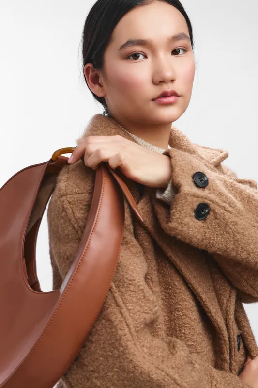 asian woman wearing a textured brown coat and carrying a large leather bag, posing against a light background. confident and stylish, she projects a modern chic look.