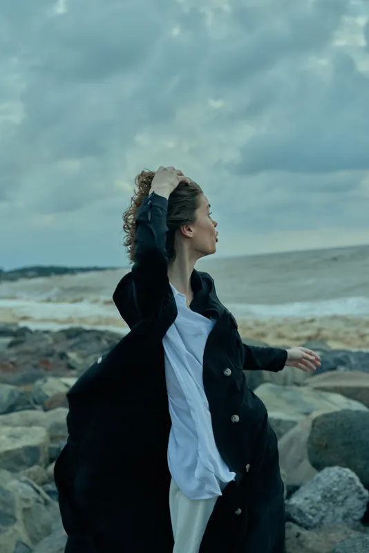 Woman on a windy coastal scene, wearing an open jacket and flowing shirt, standing on rocks with ocean and moody clouds behind her, conveying motion, freedom, and coastal styling.