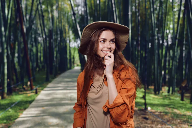 Woman smile forest path hat portrait of a happy young traveler on a tree-lined trail, wearing an orange jacket and wide brim hat, enjoying nature, relaxed outdoors lifestyle and calm scenery.
