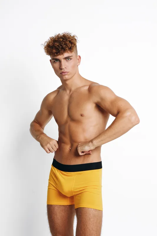 Athletic young man with curly hair posing shirtless in bright yellow boxer shorts studio shot, muscular body and confident expression, isolated white background indoor.