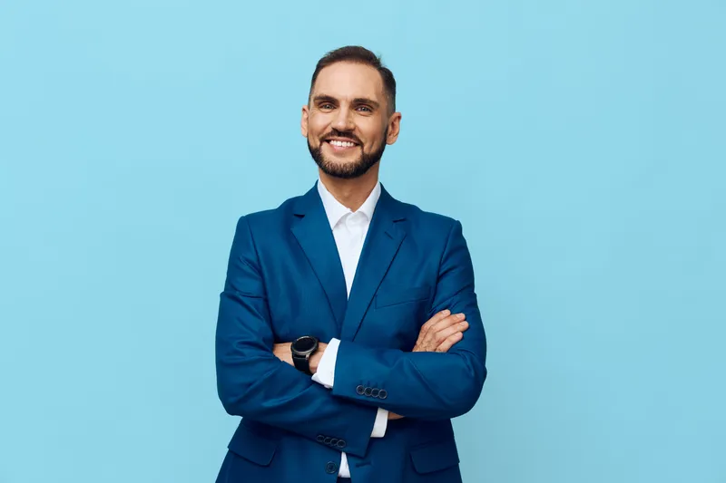 A confident professional man stands against a solid colored backdrop, dressed in a tailored suit. His posture and warm smile convey poised optimism and readiness for business challenges.