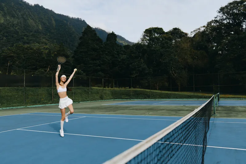Young athlete celebrating victory on a tennis court, wearing a white sporty outfit, surrounded by lush greenery and blue court, showcasing joy and energy