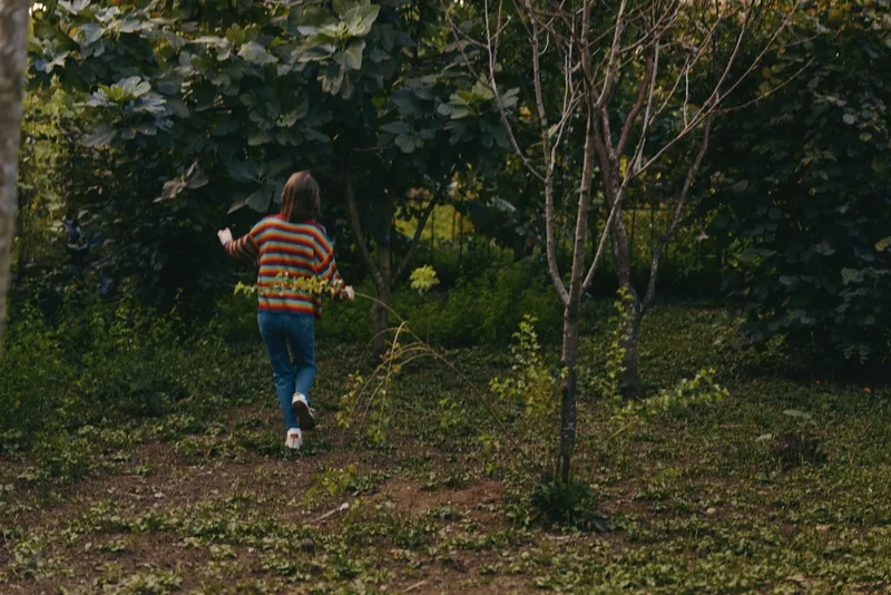 Girl in a striped sweater walking through an overgrown garden path, back view of a curious child exploring nature in a casual outfit and jeans, playful mood in soft green foliage.